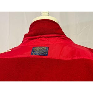 May include: A close-up of a red wool jacket, showcasing the back of the collar and upper back. A blue label with gold text is visible. The jacket is made of a thick, textured material, with a smooth, quilted lining.