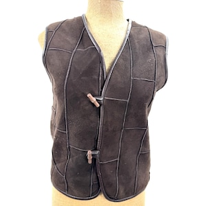 May include: A dark brown sleeveless waistcoat constructed from multiple panels of suede-like material with visible stitched seams. The garment features a V-neckline and two light brown wooden toggle closures positioned vertically along the front opening.
