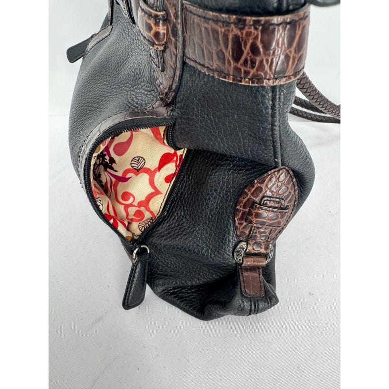 May include: Black leather handbag with brown crocodile-textured accents. The bag features a zippered pocket with a patterned red and cream lining. The bag has a shoulder strap and a decorative buckle.