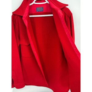 May include: A bright red wool coat with a collared neckline and two front pockets. The coat is open, revealing a red interior lining and a label. The coat is hanging on a white hanger.