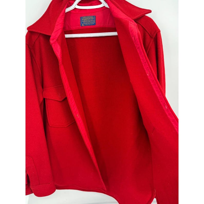 May include: A bright red wool coat with a collared neckline and two front pockets. The coat is open, revealing a red interior lining and a label. The coat is hanging on a white hanger.