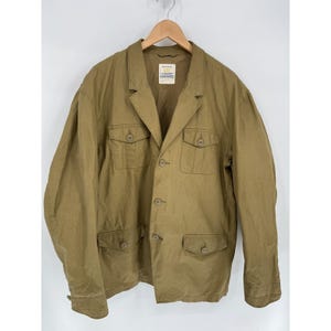 May include: A khaki-coloured blazer with four front pockets and button fastenings. The jacket has a notched lapel and is hanging on a wooden hanger. The garment has a tag on the inside collar.