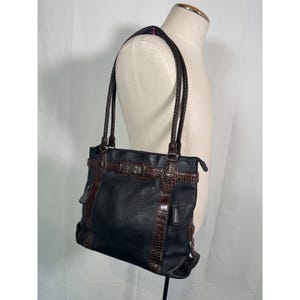 May include: Black leather tote bag with brown crocodile-textured accents and braided handles. The bag features a silver-toned buckle and a structured design. The bag is displayed on a mannequin.