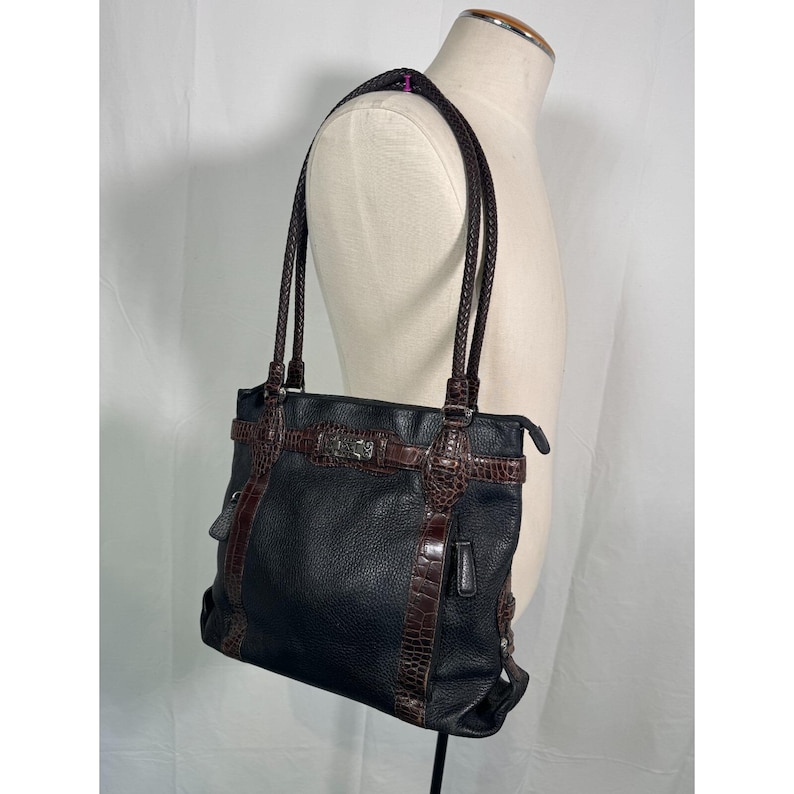 May include: Black leather tote bag with brown crocodile-textured accents and braided handles. The bag features a silver-toned buckle and a structured design. The bag is displayed on a mannequin.