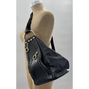 May include: A black leather hobo bag featuring a single wide shoulder strap attached to a large gold-tone metal ring. The bag includes two vertical zippered pockets on the front, each accented with gold-tone circular zipper pulls and studs.