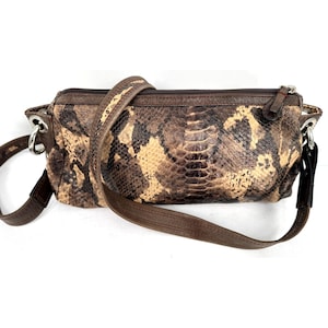 Op de afbeelding: Een bruine en tan crossbodytas met een slangenpatroon en een lange, verstelbare riem. De tas heeft een ritssluiting aan de bovenkant en zilverkleurige hardware. Het ontwerp van de tas heeft een mix van bruine en tan tinten.