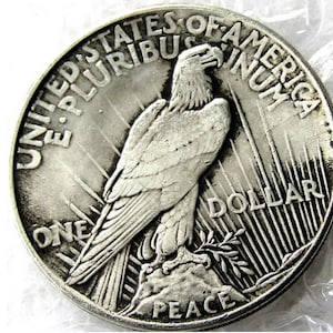 May include: A silver US Peace Dollar coin with the words "United States of America" and "One Dollar" inscribed around the edge. The coin features a bald eagle with outstretched wings, perched on a branch with the word "Peace" below.