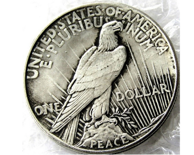 May include: A silver US Peace Dollar coin with the words "United States of America" and "One Dollar" inscribed around the edge. The coin features a bald eagle with outstretched wings, perched on a branch with the word "Peace" below.