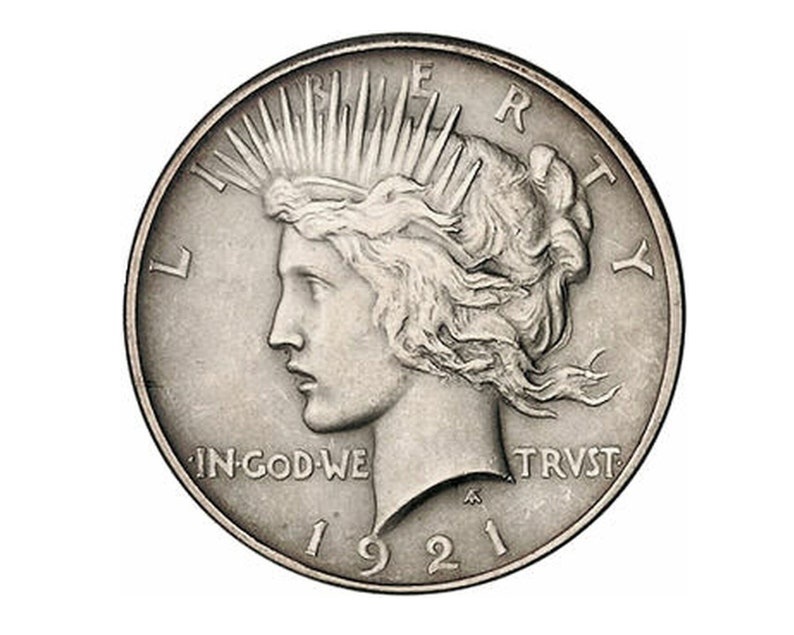 May include: A silver Peace dollar coin with the year 1921 inscribed on the bottom. The coin features a profile of a woman with flowing hair and the words "Liberty" and "In God We Trust" inscribed on the coin.