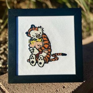 Pode incluir: Uma bordadura de ponto cruz enquadrada que apresenta um tigre de desenho animado abraçando uma pequena figura humana. O tigre é laranja e preto com listras brancas, e a figura humana está vestindo uma camisa listrada vermelha e branca. A bordadura está em tecido branco e está enquadrada em uma moldura preta.