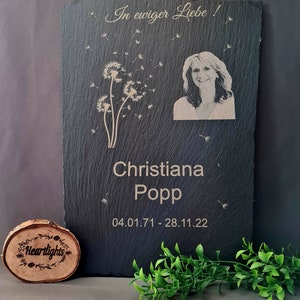 May include: A rectangular black slate memorial plaque featuring an engraved portrait of a woman, a dandelion graphic, and the text In ewiger Liebe, Christiana Popp, and the dates 04.01.71 - 28.11.22. A small circular wooden disc with the text Heartlights is included.