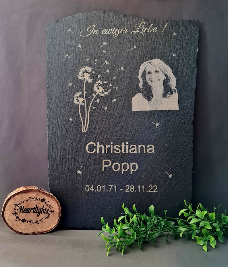 May include: A rectangular black slate memorial plaque featuring an engraved portrait of a woman, a dandelion graphic, and the text In ewiger Liebe, Christiana Popp, and the dates 04.01.71 - 28.11.22. A small circular wooden disc with the text Heartlights is included.