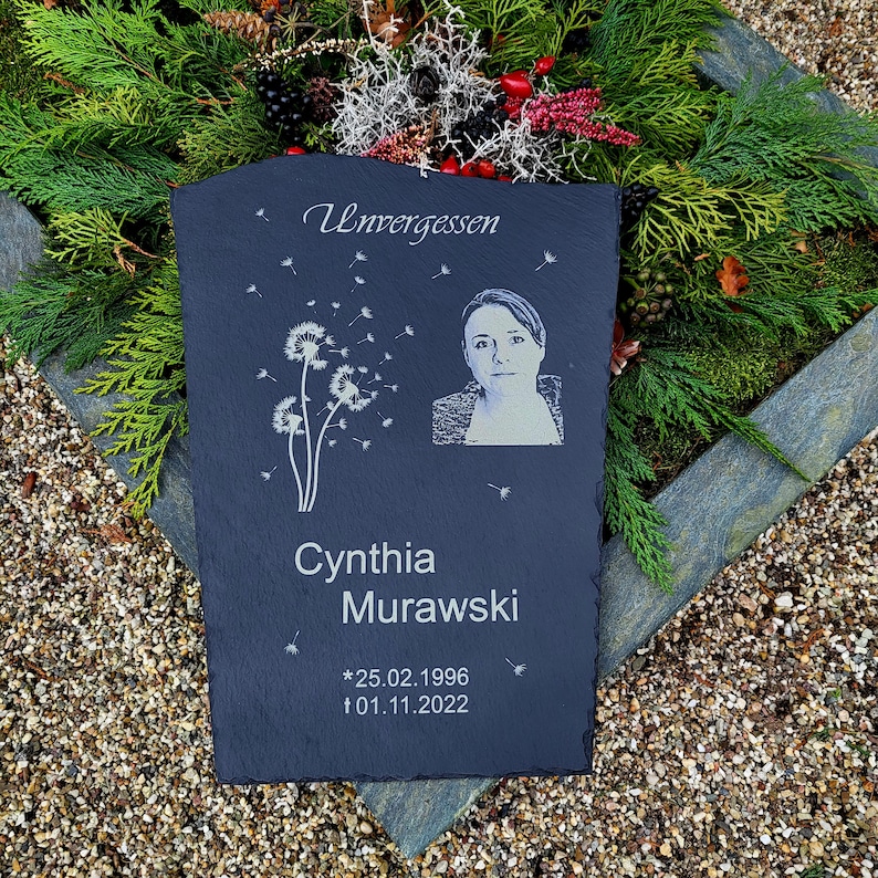 May include: A rectangular dark gray slate memorial plaque featuring the engraved text Unvergessen, the name Cynthia Murawski, and the dates 25.02.1996 and 01.11.2022. The plaque includes a laser-etched portrait of a person and dandelion seed graphics.