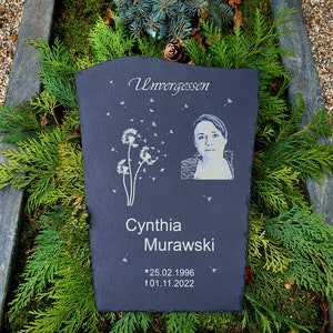 May include: A dark gray slate memorial plaque featuring the engraved text Unvergessen, a dandelion graphic with floating seeds, a portrait of a person, the name Cynthia Murawski, and the dates 25.02.1996 and 01.11.2022 with star and cross symbols.