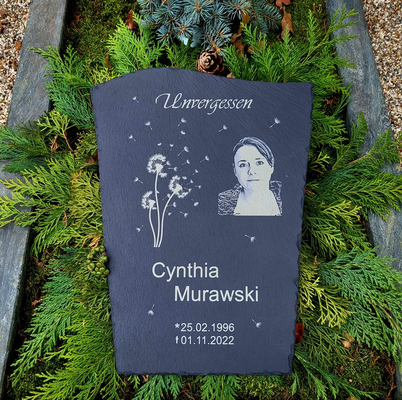 May include: A dark gray slate memorial plaque featuring the engraved text Unvergessen, a dandelion graphic with floating seeds, a portrait of a person, the name Cynthia Murawski, and the dates 25.02.1996 and 01.11.2022 with star and cross symbols.