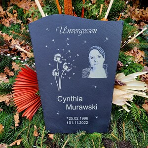 May include: A dark gray slate memorial plaque featuring an engraved portrait of a person, a dandelion graphic, and the text Unvergessen, Cynthia Murawski, *25.02.1996, and &dagger;01.11.2022. The plaque has a textured, uneven top edge and a smooth surface.