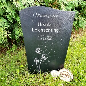 May include: A dark gray slate memorial plaque with a tapered shape, engraved with the text Unvergessen, Ursula Leichsenring, and the dates 17.01.1945 and 16.03.2018. The plaque features a white dandelion graphic with seeds blowing in the wind.