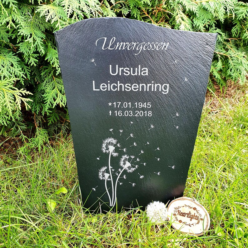 May include: A dark gray slate memorial plaque with a tapered shape, engraved with the text Unvergessen, Ursula Leichsenring, and the dates 17.01.1945 and 16.03.2018. The plaque features a white dandelion graphic with seeds blowing in the wind.