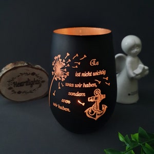 May include: A matte black cylindrical glass candle holder featuring a glowing orange cutout design of a dandelion, an anchor, and the German text: Es ist nicht wichtig was wir haben, sondern wen wir haben. The interior glows with warm light.