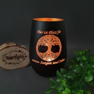 May include: A black cylindrical candle holder with a warm orange interior glow. The exterior features a laser-cut tree of life design and the German text "Hier ist Platz für meine Sorgen und Nöte" illuminated by light from within the vessel.