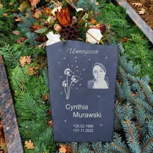 May include: A rectangular slate memorial plaque featuring the engraved text Unvergessen, the name Cynthia Murawski, and the dates 25.02.1996 and 01.11.2022. The plaque includes an etched portrait of a person and a graphic of dandelion seeds.