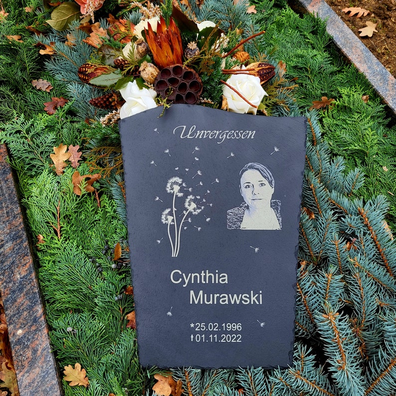 May include: A rectangular slate memorial plaque featuring the engraved text Unvergessen, the name Cynthia Murawski, and the dates 25.02.1996 and 01.11.2022. The plaque includes an etched portrait of a person and a graphic of dandelion seeds.