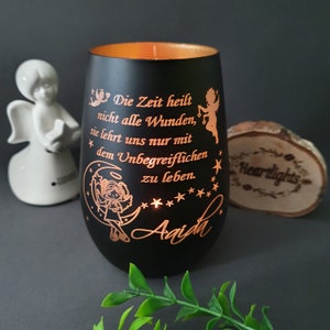 May include: A black cylindrical candle holder with a metallic gold interior. The exterior features engraved copper-colored text reading "Die Zeit heilt nicht alle Wunden, sie lehrt uns nur mit dem Unbegreiflichen zu leben." and the name "Aaida" with angel and moon graphics.