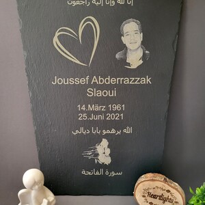 May include: A black slate memorial plaque featuring an engraved portrait of a man, a heart outline, the name Joussef Abderrazzak Slaoui, dates 14.M&auml;rz 1961 and 25.Juni 2021, Arabic text, and a rose illustration, accompanied by a small white angel figurine.