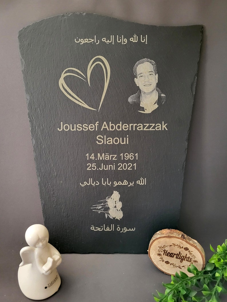 May include: A black slate memorial plaque featuring an engraved portrait of a man, a heart outline, the name Joussef Abderrazzak Slaoui, dates 14.M&auml;rz 1961 and 25.Juni 2021, Arabic text, and a rose illustration, accompanied by a small white angel figurine.