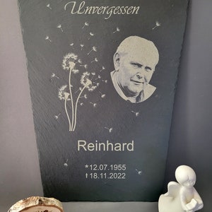 May include: A rectangular slate memorial plaque featuring an engraved portrait of a man, a dandelion flower design, and the text Unvergessen, Reinhard, 12.07.1955, and 18.11.2022. Beside it sits a small white angel figurine and a wooden disc with Heartlights.