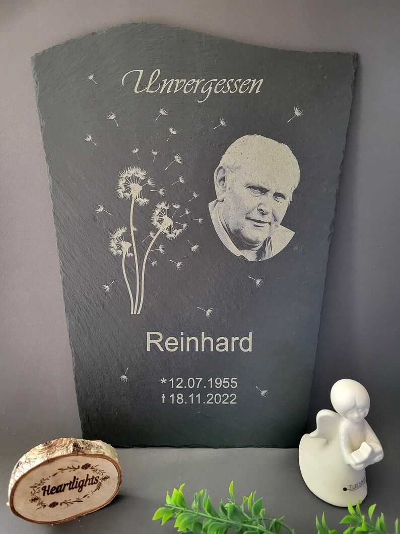 May include: A rectangular slate memorial plaque featuring an engraved portrait of a man, a dandelion flower design, and the text Unvergessen, Reinhard, 12.07.1955, and 18.11.2022. Beside it sits a small white angel figurine and a wooden disc with Heartlights.