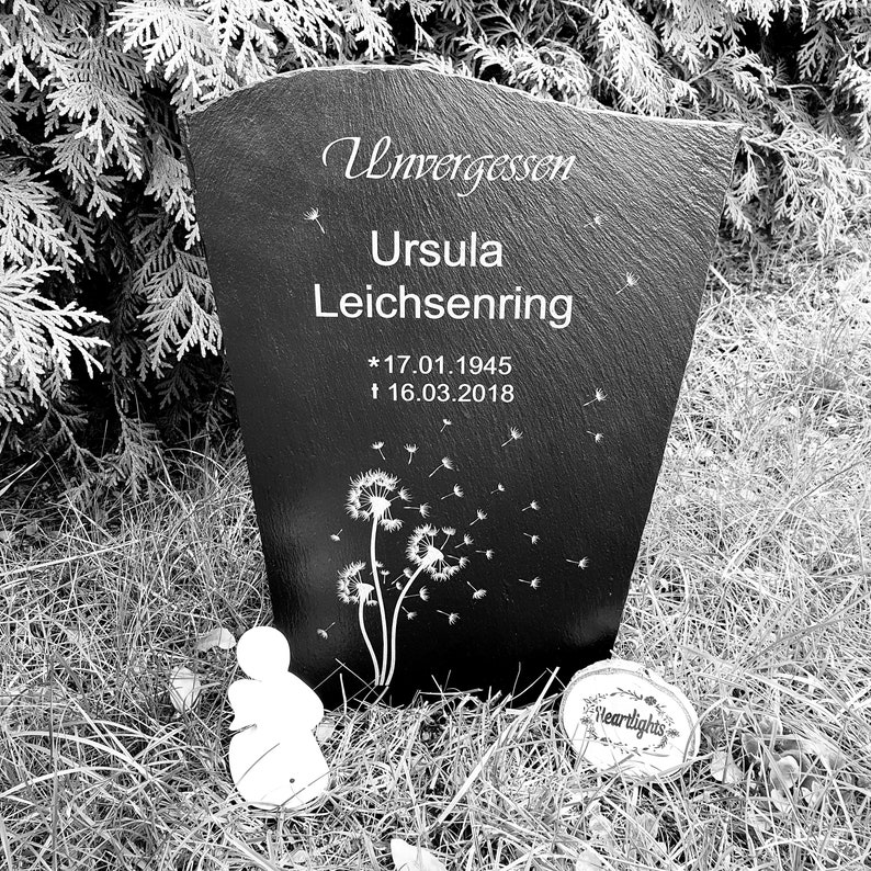 May include: A black slate memorial plaque featuring the engraved text Unvergessen, Ursula Leichsenring, and the dates 17.01.1945 and 16.03.2018. The surface is decorated with white dandelion illustrations and floating seeds, accompanied by a small angel figurine.