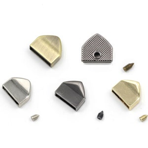 Könnte beinhalten: Vier Metallteile für Taschen oder Handtaschen in verschiedenen Ausführungen: Antikmessing, Silber, Schwarz und Gold. Jedes Teil hat eine dreieckige Form mit einem kleinen Loch in der Mitte. Die Teile sind auf einer weißen Oberfläche mit kleinen Schrauben daneben angeordnet.