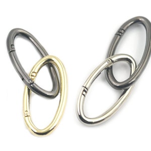 May include: Two oval-shaped metal clasps, one in gold and one in black, are linked together. The clasps are designed to be used as a connector for bags or other items.