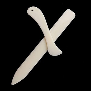 Puede incluir: Dos herramientas de papel de hueso blanco, una es una hoja larga y delgada y la otra es una hoja curva con un agujero en la parte superior.