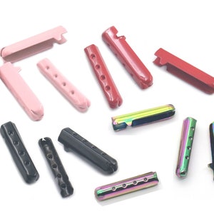 May include: Assortment of metal cord ends in various colors, including pink, red, black, and iridescent. The cord ends have multiple holes for threading cords or strings.