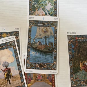 May include: A collection of tarot cards featuring detailed illustrations. The cards display various scenes, including a boat on water, figures in a garden, and a rider on horseback. Text in multiple languages is visible on the cards.