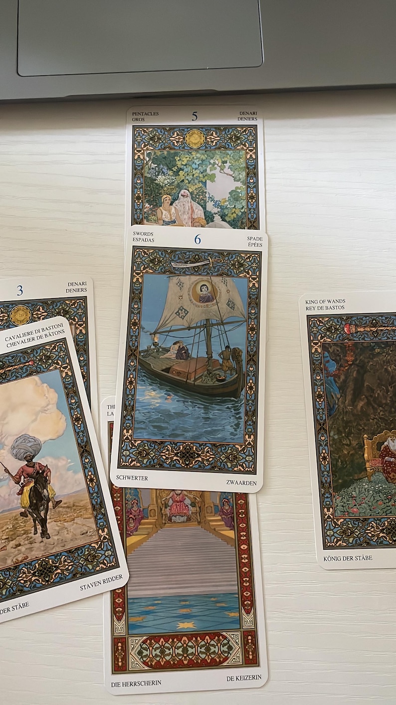 May include: A collection of tarot cards featuring detailed illustrations. The cards display various scenes, including a boat on water, figures in a garden, and a rider on horseback. Text in multiple languages is visible on the cards.