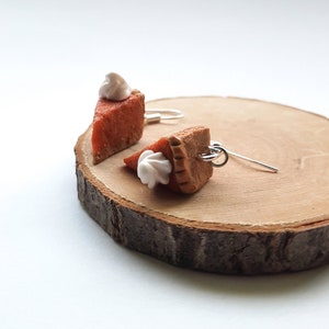 Könnte beinhalten: Ein Paar Hängeohrringe mit Miniatur-Kürbiskuchenstücken mit weißem Frosting. Die Ohrringe werden auf einer Holzscheibe präsentiert.