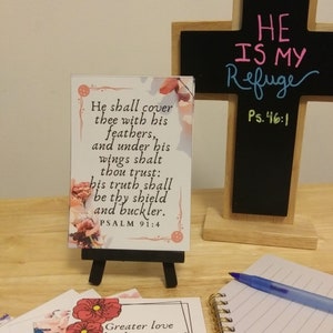 May include: A small wooden easel displaying a white card with a pink floral design and a bible verse: "He shall cover thee with his feathers, and under his wings shalt thou trust: his truth shall be thy shield and buckler. PSALM 91:4". A black chalkboard cross with the words "HE IS MY Refuge Ps. 46:1" is in the background.