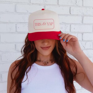 May include: A cream-colored baseball cap with a red brim and top button. The cap features the text "THIS & YAP" in a red rectangular patch. The hat is worn by a person in a white tank top.