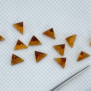 May include: A collection of twelve triangular-shaped, faceted, amber-colored gemstones. The gemstones are arranged on a white surface.