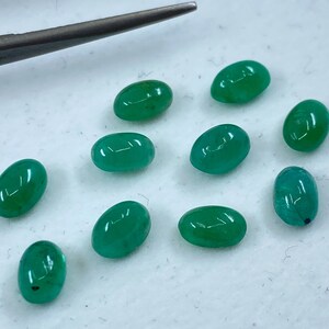 May include: Ten oval-shaped green gemstones, likely emeralds, are scattered on a white surface. The gemstones are smooth and polished, with a slight variation in size and shape.