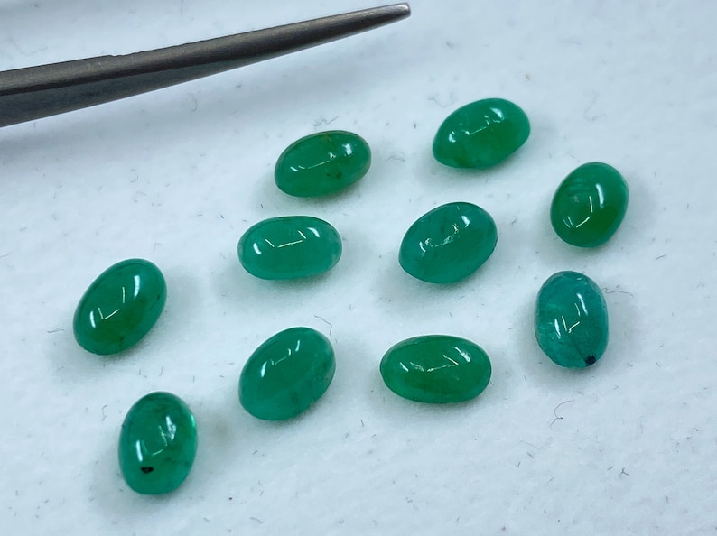 May include: Ten oval-shaped green gemstones, likely emeralds, are scattered on a white surface. The gemstones are smooth and polished, with a slight variation in size and shape.