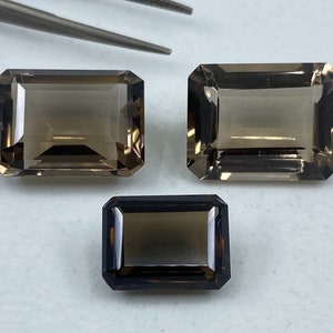 May include: Three smoky quartz gemstones in emerald cut. The gemstones are different sizes and are arranged in a row on a white surface.