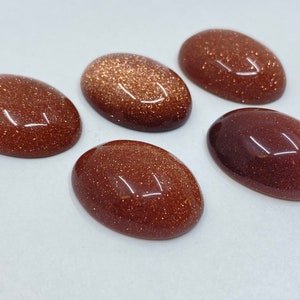 May include: Five oval-shaped, polished gemstones in varying shades of brown and reddish-brown. The stones have a sparkling, glittery appearance, with a smooth, reflective surface. The gemstones are arranged on a white surface.