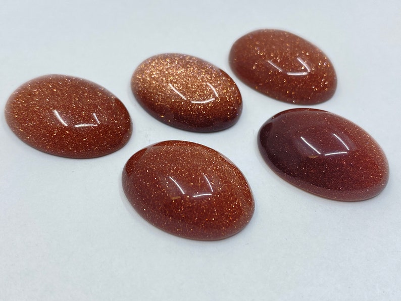 May include: Five oval-shaped, polished gemstones in varying shades of brown and reddish-brown. The stones have a sparkling, glittery appearance, with a smooth, reflective surface. The gemstones are arranged on a white surface.