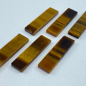 May include: Six polished tiger's eye stones, each cut into a rectangular shape. The stones are a rich golden brown colour with a subtle shimmer.