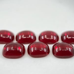 May include: Seven red cabochon gemstones, each with a smooth, rounded surface, arranged in a row on a white background.