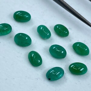 May include: Ten oval-shaped green gemstones, likely emeralds, are arranged on a white surface. A metal tool is visible in the background.